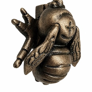 Puede incluir: Un llamador de puerta de metal en tono bronce con forma de abeja. El insecto presenta una cabeza redondeada con una marca en forma de cruz, un cuerpo segmentado y alas texturizadas extendidas hacia los lados. Incluye un mecanismo articulado.