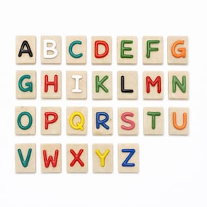 Puede incluir: Azulejos del alfabeto con letras coloridas. Cada azulejo presenta una letra diferente del alfabeto en una variedad de colores, incluyendo negro, blanco, rojo, verde y naranja. Los azulejos están dispuestos sobre una superficie blanca.