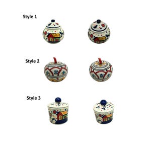 May include: Three styles of ceramic storage jars with lids. Style 1 is a round jar with a house design. Style 2 is a round jar with a floral design. Style 3 is a cylindrical jar with a house design.