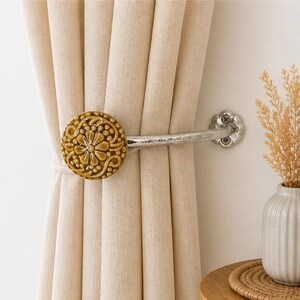 May include: A beige curtain held back by a metal tieback featuring a round, mustard-yellow ceramic knob with a raised floral and scroll pattern. The tieback is secured to the wall with a silver-toned, flower-shaped metal mounting plate.