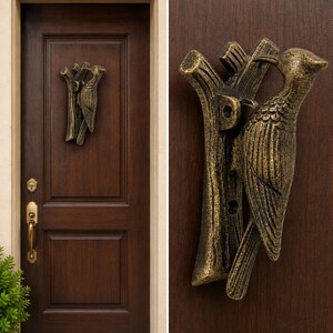 May include: A bronze-toned metal door knocker shaped like a woodpecker perched on a tree branch. The bird features detailed wing feathers and a long beak, mounted vertically on a dark brown wooden door with a matching brass-colored handle below.