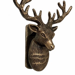 Puede incluir: Un gancho de pared de metal en tono bronce con forma de cabeza de ciervo, con astas detalladas y pelaje texturizado. El gancho cuenta con una placa de montaje vertical unida a la parte posterior de la cabeza para colgar objetos.