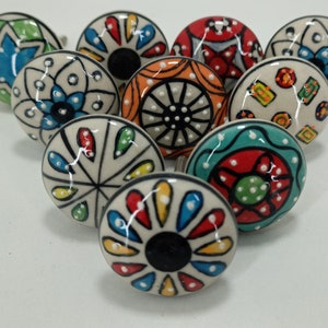 Op de afbeelding: Een verzameling van tien keramische kastknoppen. Elke knop heeft een uniek handgeschilderd ontwerp met een witte basis en kleurrijke patronen. De ontwerpen omvatten bloemen, geometrische en abstracte motieven in verschillende kleuren.