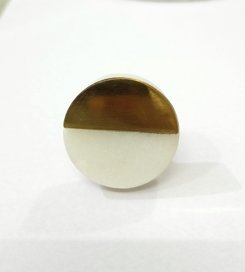 K&ouml;nnte beinhalten: Ein runder Schrankknopf mit einem Halb-und-Halb-Design. Die obere H&auml;lfte ist goldfarben geb&uuml;rstet, die untere H&auml;lfte ist wei&szlig;. Der Knopf besteht aus Metall und Marmor.