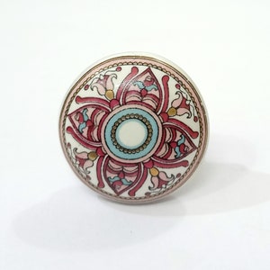 May include: A round ceramic knob with a pink, blue, and gold floral design. The knob has a white background and a gold rim.
