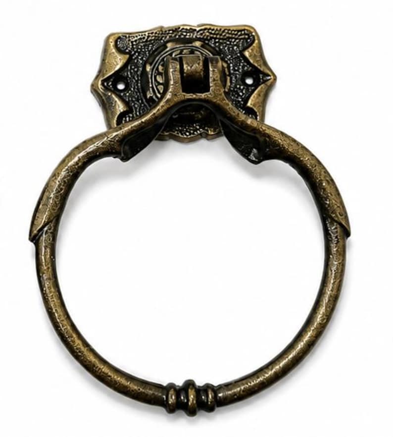 Puede incluir: Un tirador de anillo de metal en tono lat&oacute;n antiguo con un acabado texturizado y desgastado. El anillo circular est&aacute; unido a una placa de montaje rectangular decorativa con detalles en relieve y dos orificios visibles para tornillos.