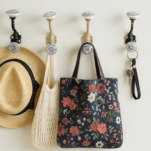 May include: Four decorative wall hooks with floral ceramic knobs. A straw hat with a black band, a white net bag, a navy tote bag with a red floral pattern, and a set of keys hang from the hooks. The hooks are black and gold.