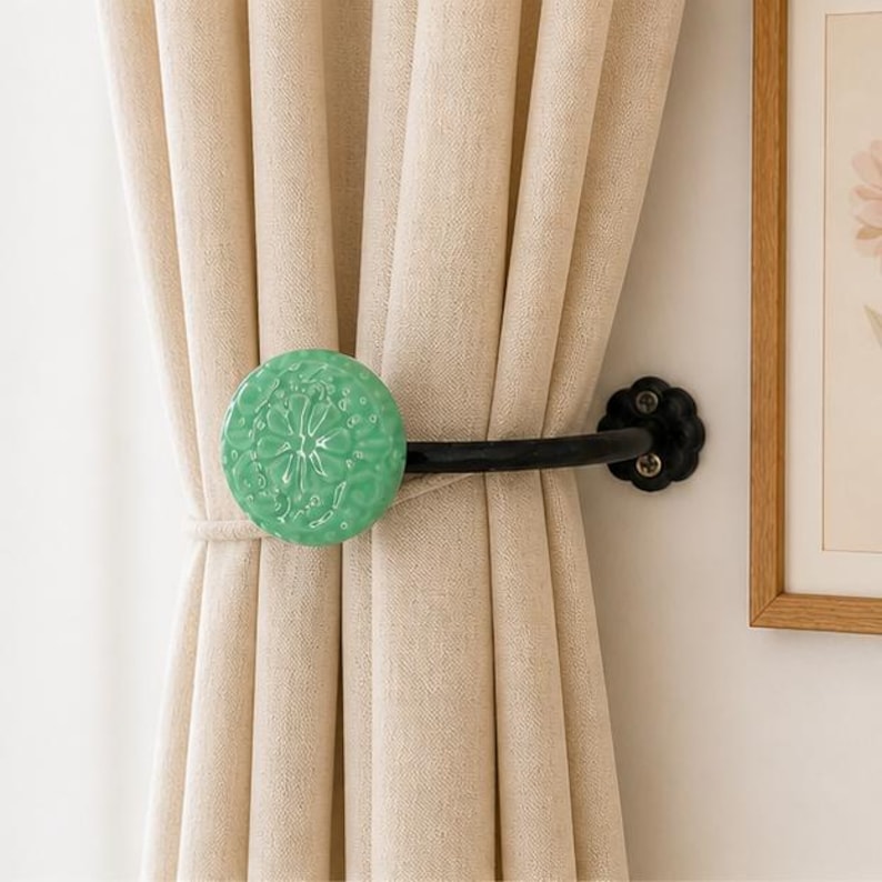 May include: A curtain tieback featuring a round, mint green ceramic knob with a raised floral pattern. The knob is attached to a curved, matte black metal arm that mounts to the wall with a scalloped black base plate, holding beige fabric curtains.