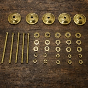 May include: A set of brass hardware components, including five circular mounting plates, long threaded bolts, washers, and nuts. The hardware is arranged on a dark wooden surface, showcasing the various pieces for assembly.