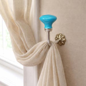 May include: A wall-mounted curtain tieback hook featuring a bright turquoise ceramic knob with a smooth, glossy finish. The hook has a polished silver-tone metal arm and a decorative, scalloped circular base plate attached to the wall surface.