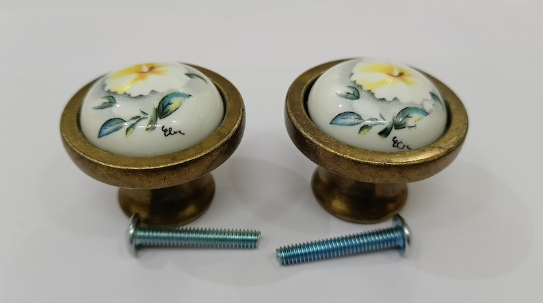 Pu&ograve; includere: Due pomelli per armadi in ottone con top in ceramica bianca che presentano un fiore giallo con foglie verdi. Ogni pomo viene fornito con una vite argentata.