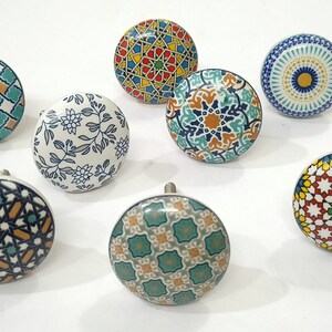 Może przedstawiać: Kolekcja dziewięciu dekoracyjnych ceramicznych gałek do szafek. Gałki mają r&oacute;żne kolorowe geometryczne i kwiatowe wzory w odcieniach niebieskiego, zielonego, ż&oacute;łtego i pomarańczowego. Każda gałka ma srebrną metalową podstawę.