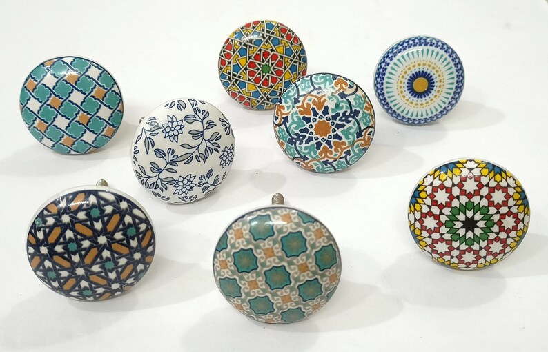 Może przedstawiać: Kolekcja dziewięciu dekoracyjnych ceramicznych gałek do szafek. Gałki mają r&oacute;żne kolorowe geometryczne i kwiatowe wzory w odcieniach niebieskiego, zielonego, ż&oacute;łtego i pomarańczowego. Każda gałka ma srebrną metalową podstawę.