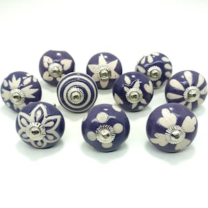May include: A set of ten decorative ceramic cabinet knobs. The knobs are a deep purple colour with white patterns, including floral, polka dot, and striped designs. Each knob has a silver-coloured metal base.