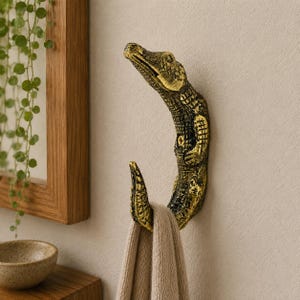 Peut inclure: Un crochet mural en forme d'alligator avec une finition métallique dorée et noire texturée. Le corps de l'alligator se courbe pour former le crochet, avec sa queue pointant vers le haut. Une serviette en tissu beige est suspendue à la queue.