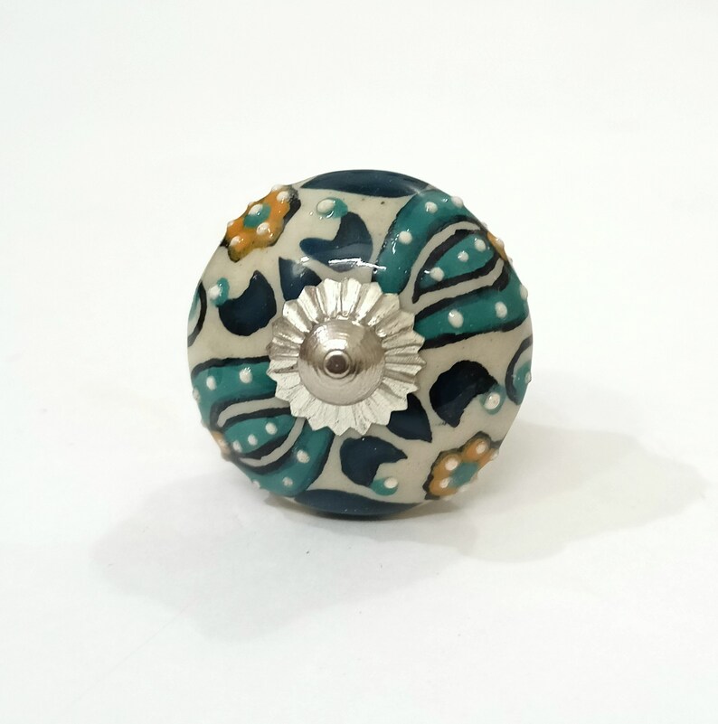 Op de afbeelding: Een witte keramische knop met een blauw en groen bloemenpatroon en een zilveren metalen midden.