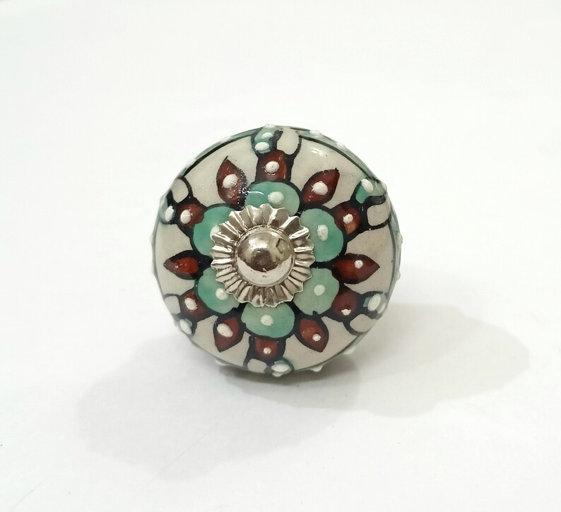 Op de afbeelding: Een keramische knop met een witte achtergrond en een bloemmotief in tinten groen, bruin en wit. De knop heeft een zilveren metalen midden.