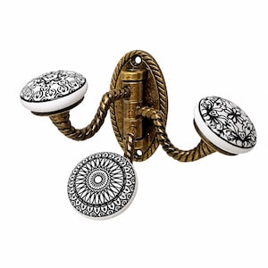 Può includere: Un gancio triplo da parete con una base ovale in ottone anticato dal design a corda intrecciata. Tre bracci curvi si estendono dal centro, ciascuno sormontato da un pomello in ceramica bianca decorato con intricati motivi floreali neri.