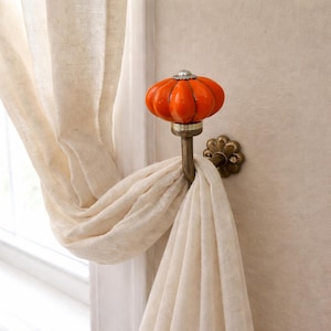 May include: A decorative curtain tieback featuring an orange ceramic pumpkin with a silver cap. The tieback is mounted on a wall and holds a cream-coloured curtain. The hardware is brass. Suitable for autumn decor, the pumpkin measures approximately 7 cm in diameter.
