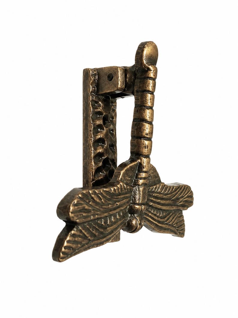 Puede incluir: Un llamador de puerta de metal en tono bronce con forma de lib&eacute;lula, alas texturizadas y cuerpo segmentado. La lib&eacute;lula est&aacute; unida a una placa de montaje vertical rectangular que presenta un acabado de superficie martillado y r&uacute;stico.