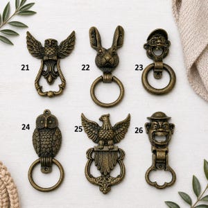 May include: A selection of six antique bronze-coloured door knockers. Designs include an owl, a rabbit, a man with a hat, an owl, an eagle, and a man with a hat. Each knocker has a ring for knocking.