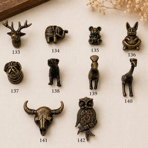 Op de afbeelding: Een collectie van tien bronskleurige metalen knoppen of ladehandgrepen in diervorm, genummerd van 133 tot 142. De figuren omvatten een hert, olifant, beer, konijn, mopshond, hond, lama, giraf, stierenschedel en een uil met gedetailleerde texturen.