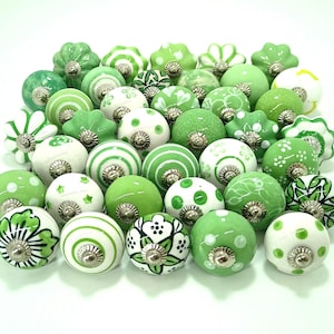 May include: A collection of green and white ceramic cabinet knobs. The knobs feature various designs, including stripes, polka dots, floral patterns, and star shapes. Each knob has a silver-colored metal base.