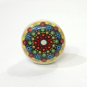 May include: A round ceramic knob featuring a colourful mandala design. The mandala has a central white circle surrounded by concentric circles of red, orange, yellow, green, blue and black dots.