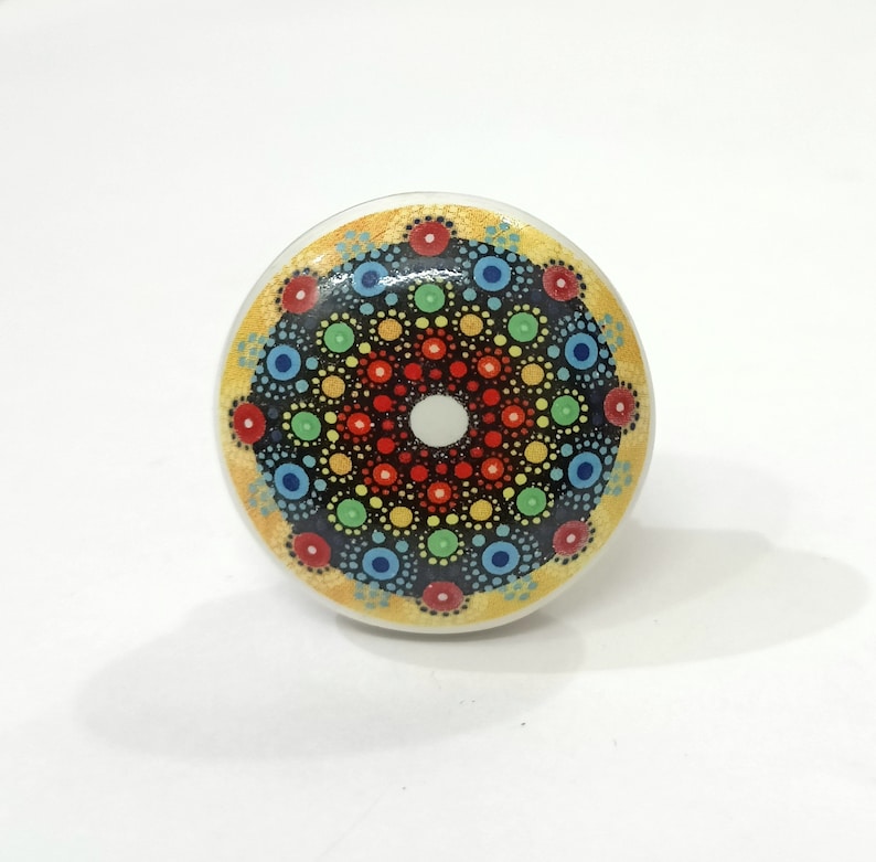 May include: A round ceramic knob featuring a colourful mandala design. The mandala has a central white circle surrounded by concentric circles of red, orange, yellow, green, blue and black dots.