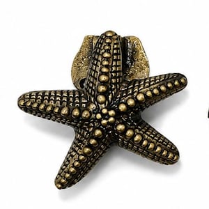 Puede incluir: Un tirador de mueble de metal en tono lat&oacute;n con forma de estrella de mar de cinco puntas. La superficie presenta un patr&oacute;n texturizado de puntos en relieve en cada brazo, con un acabado antiguo que resalta los detalles del animal marino.