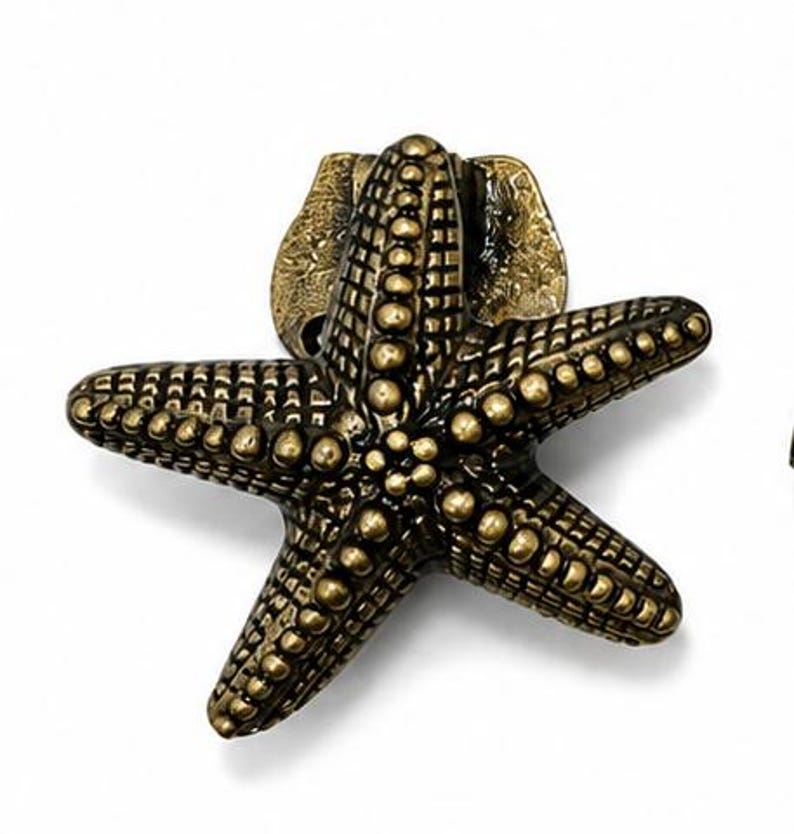 Puede incluir: Un tirador de mueble de metal en tono lat&oacute;n con forma de estrella de mar de cinco puntas. La superficie presenta un patr&oacute;n texturizado de puntos en relieve en cada brazo, con un acabado antiguo que resalta los detalles del animal marino.