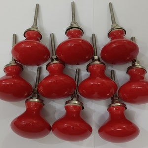 K&ouml;nnte beinhalten: Zehn rote Keramik-Schrankkn&ouml;pfe mit silbernen Metallstielen. Die Kn&ouml;pfe sind rund und haben eine leicht erh&ouml;hte Mitte.