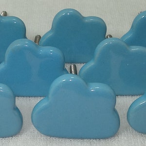 Pode incluir: Uma cole&ccedil;&atilde;o de dez puxadores de arm&aacute;rio em forma de nuvem azul clara. Cada puxador tem uma superf&iacute;cie lisa e brilhante e um pino de metal prateado para fixa&ccedil;&atilde;o. Os puxadores est&atilde;o dispostos sobre uma superf&iacute;cie branca.