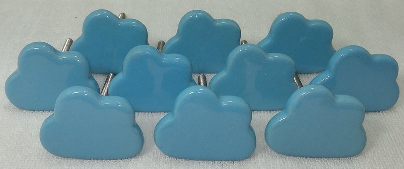 Pode incluir: Uma cole&ccedil;&atilde;o de dez puxadores de arm&aacute;rio em forma de nuvem azul clara. Cada puxador tem uma superf&iacute;cie lisa e brilhante e um pino de metal prateado para fixa&ccedil;&atilde;o. Os puxadores est&atilde;o dispostos sobre uma superf&iacute;cie branca.