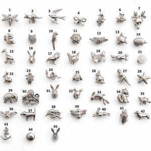 Puede incluir: Una colección de pequeños dijes metálicos plateados en varias formas de animales y objetos. Los dijes incluyen una libélula, un dinosaurio, un pájaro, un lazo, un caballito de mar, una abeja, un ciervo, un oso, un gato, un loro, una flor, alas, una tortuga, un erizo, una jirafa, un conejo, un león, un zorro, una mariposa, una estrella de mar, un botón, un perro, una ardilla, un pulpo, un conejo, un delfín, un búho, un perro, un ancla y una esfera.