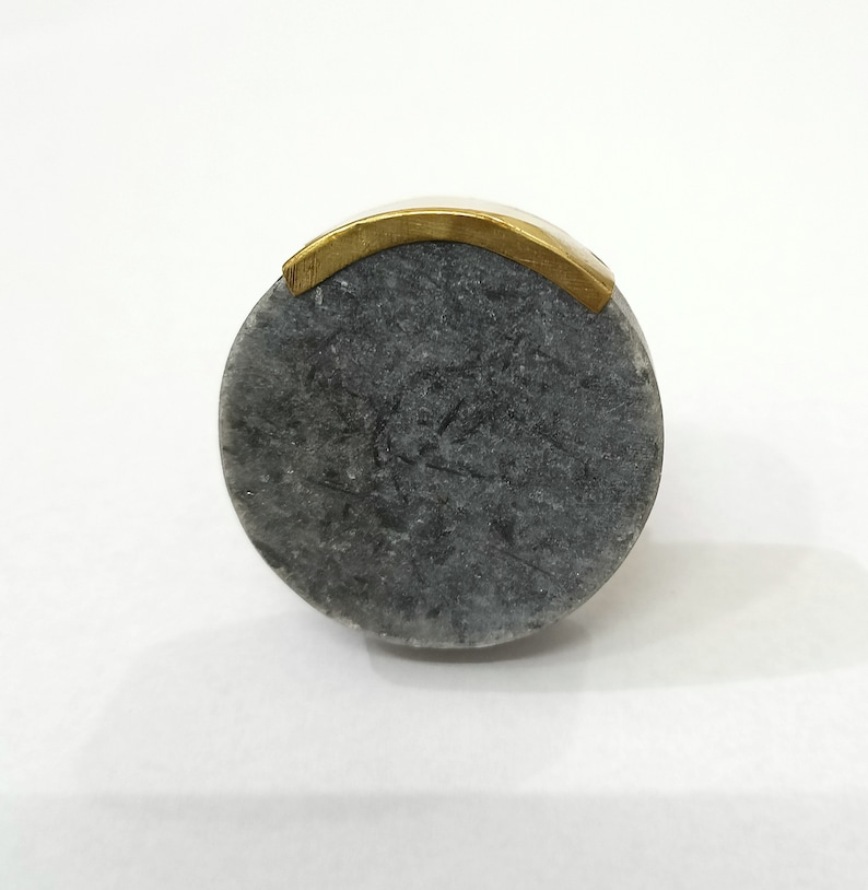 K&ouml;nnte beinhalten: Ein runder, grauer Steinring mit einem goldfarbenen Band oben. Der Ring hat ein glattes, poliertes Aussehen. Der Stein ist dunkelgrau.