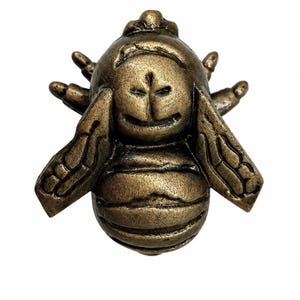 Puede incluir: Una figura de abeja de metal en tono bronce con un cuerpo redondeado y texturizado, alas detalladas que se extienden hacia los lados y una cabeza estilizada con rasgos faciales tallados. La superficie tiene un acabado met&aacute;lico con reflejos oscuros.