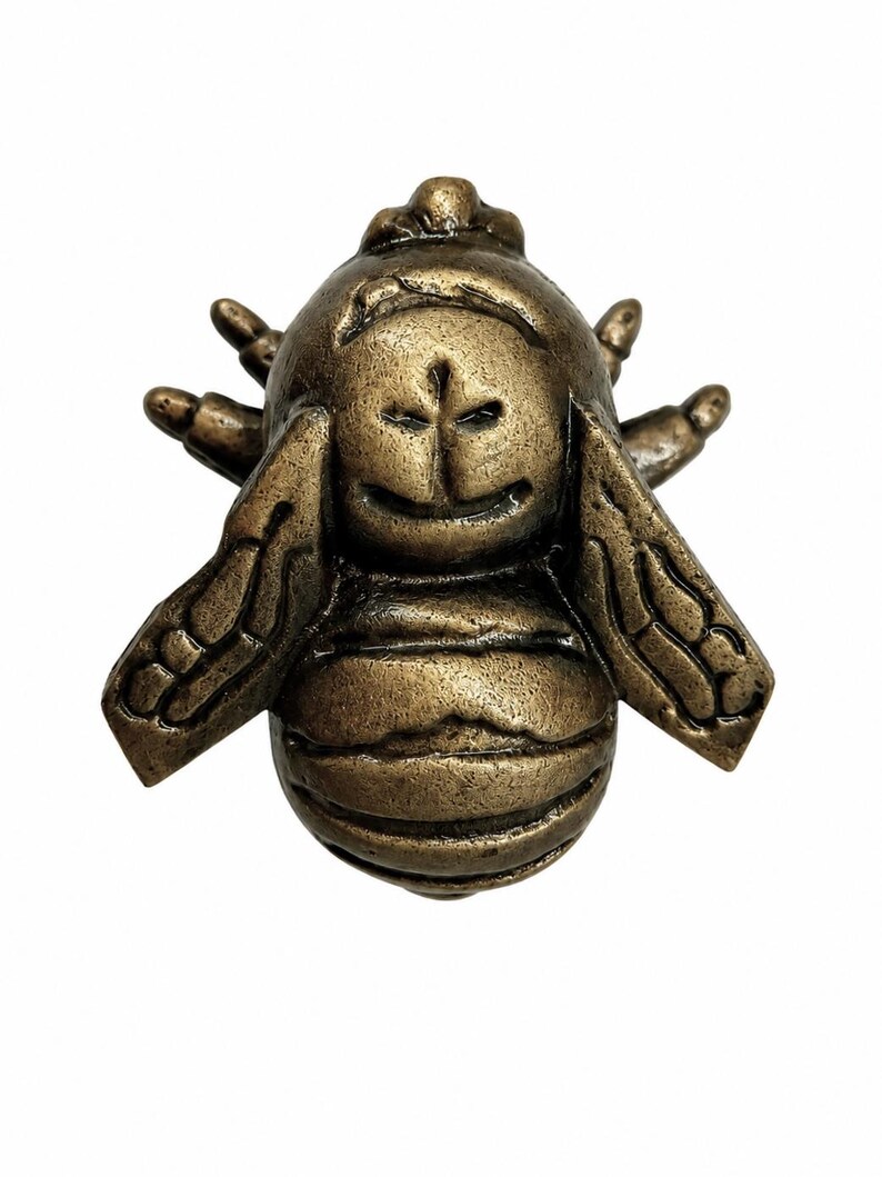 Puede incluir: Una figura de abeja de metal en tono bronce con un cuerpo redondeado y texturizado, alas detalladas que se extienden hacia los lados y una cabeza estilizada con rasgos faciales tallados. La superficie tiene un acabado met&aacute;lico con reflejos oscuros.