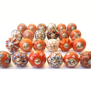 May include: A selection of orange ceramic cabinet knobs with diverse patterns. Designs include floral, geometric, and starburst motifs. Each knob has a silver-toned metal base, arranged on a white surface.