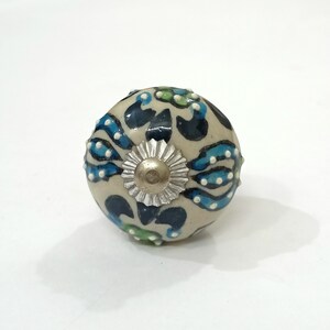 Op de afbeelding: Een witte keramische knop met een blauw, groen en zilveren bloemenontwerp. De knop is rond en heeft een glad oppervlak.