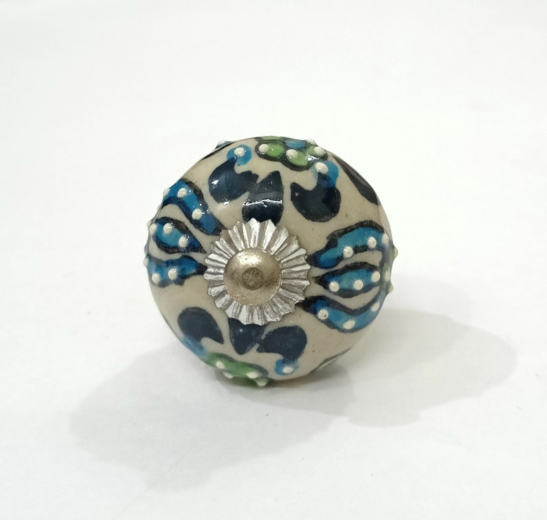 Op de afbeelding: Een witte keramische knop met een blauw, groen en zilveren bloemenontwerp. De knop is rond en heeft een glad oppervlak.