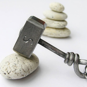Pode incluir: Um martelo de metal prateado com uma alça de nó e um chaveiro. O martelo está apoiado em uma rocha branca.