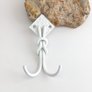 May include: White metal double hook with a knot design, mounted on a white diamond-shaped plate. The hook is painted white and has a textured surface. A rough, light-coloured stone is positioned above the hook.