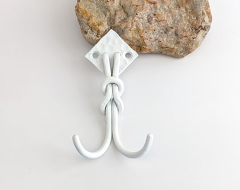 Puede incluir: Gancho doble de metal blanco con diseño de nudo, montado sobre una placa blanca en forma de diamante. El gancho está pintado de blanco y tiene una superficie texturizada. Una piedra rugosa de color claro está colocada encima del gancho.