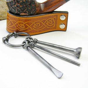 May include: A set of three metal pipe tools with a ring for attaching to a keychain. The tools are made of silver metal and have different shapes, including a round tip, a pointed tip, and a flat tip. The tools are attached to a leather strap with a Celtic knot design.
