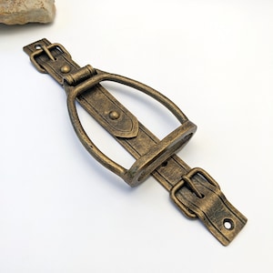 May include: A gold-toned metal wall mount with a stirrup design and two buckles. The mount is designed to hold a bottle of wine.