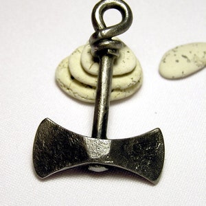 May include: A silver double-headed axe pendant with a knotted loop at the top. The pendant is resting on a stack of white stones.