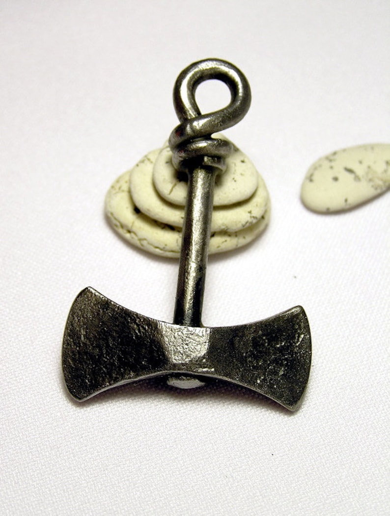 May include: A silver double-headed axe pendant with a knotted loop at the top. The pendant is resting on a stack of white stones.