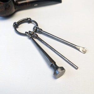 Puede incluir: Un juego de herramientas para pipa de plata con tres herramientas unidas a un anillo. Las herramientas incluyen un tampón, un escariador y un raspador.