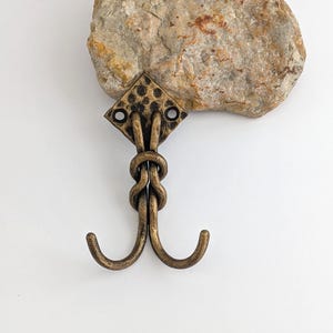 May include: Antique bronze-colored double hook with a diamond-shaped top and two curved hooks. The hook is attached to a light-colored rock. The hook is designed for hanging items.
